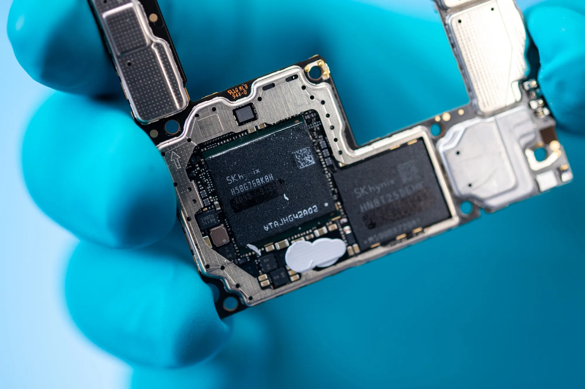 A gloved hand holding a smartphone circuit board with SK Hynix memory chips