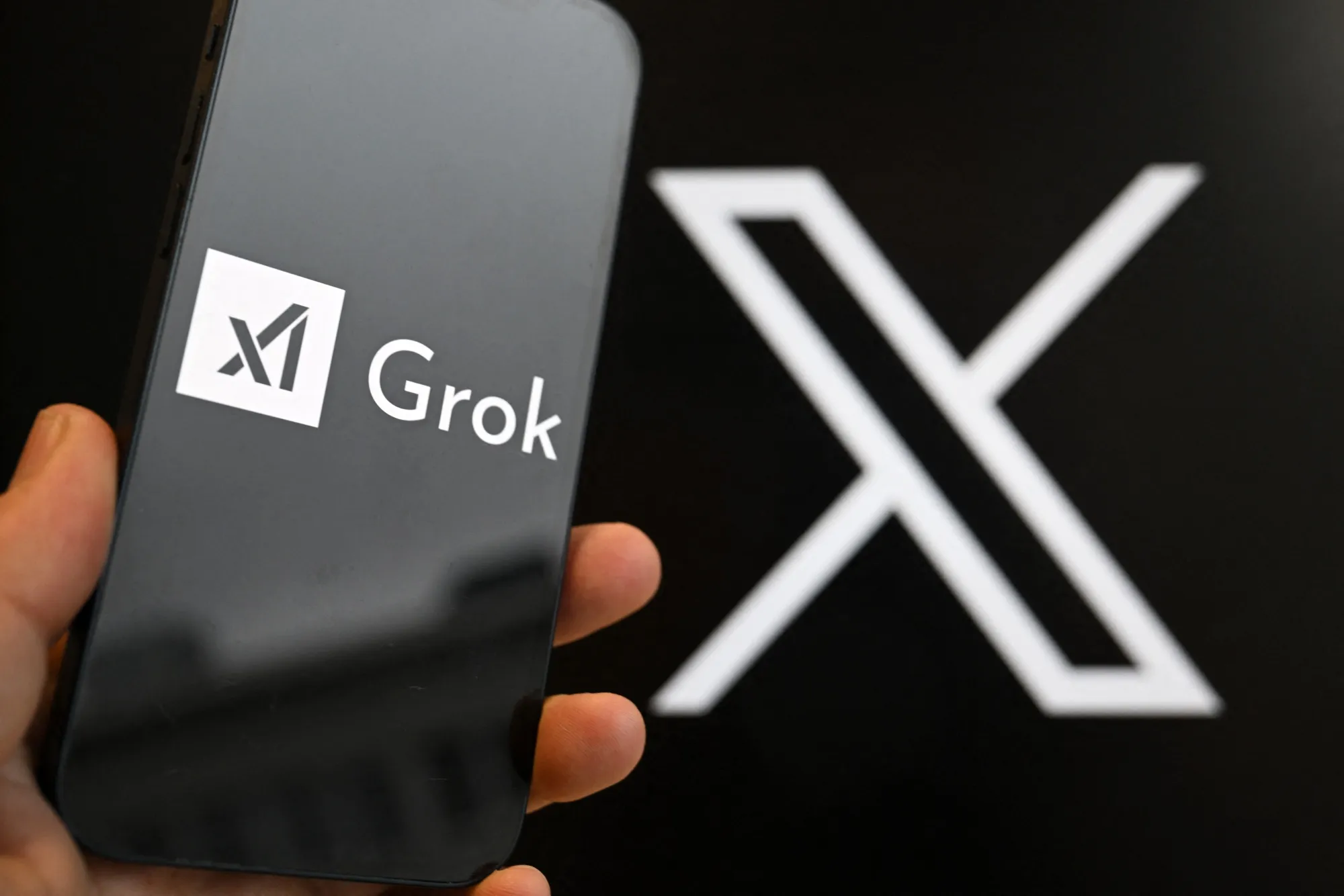 A hand holding a phone displaying the Grok app logo in front of the X logo